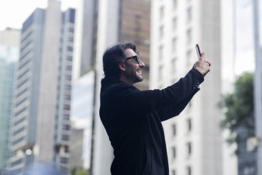 Gündüz vakti güneş gözlüklü ve akıllı telefonlu arka planda şehir manzaralı iyi giyimli genç bir adamın görüntüsü. Akıllı telefonla selfie çekiyorum..