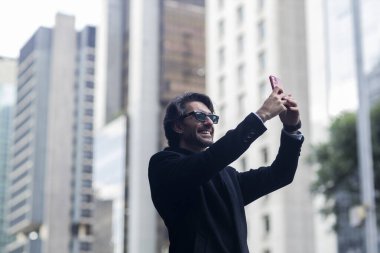 Gündüz vakti güneş gözlüklü ve akıllı telefonlu arka planda şehir manzaralı iyi giyimli genç bir adamın görüntüsü. Akıllı telefonla selfie çekiyorum..