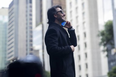 Gündüz vakti güneş gözlüklü ve akıllı telefonlu arka planda şehir manzaralı iyi giyimli genç bir adamın görüntüsü. Yüksek kalite fotoğraf. 