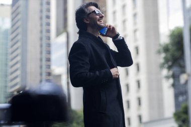 Gündüz vakti güneş gözlüklü ve akıllı telefonlu arka planda şehir manzaralı iyi giyimli genç bir adamın görüntüsü. Yüksek kalite fotoğraf. 