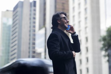 Gündüz vakti güneş gözlüklü ve akıllı telefonlu arka planda şehir manzaralı iyi giyimli genç bir adamın görüntüsü. Yüksek kalite fotoğraf. 