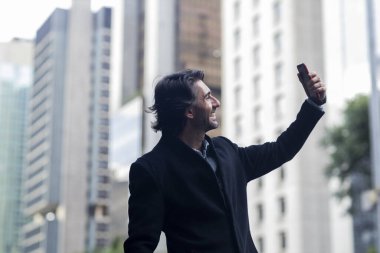 Gündüz vakti akıllı telefonlu, iyi giyimli ve arka planda şehir manzaralı genç bir adamın görüntüsü. Akıllı telefonla selfie çekiyorum..