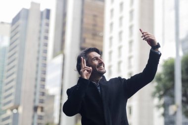 Gündüz vakti akıllı telefonlu, iyi giyimli ve arka planda şehir manzaralı genç bir adamın görüntüsü. Akıllı telefonla selfie çekiyorum..