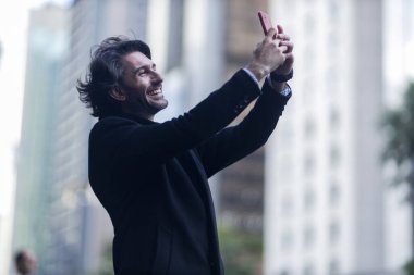 Gündüz vakti akıllı telefonlu, iyi giyimli ve arka planda şehir manzaralı genç bir adamın görüntüsü. Akıllı telefonla selfie çekiyorum..