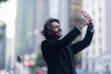 Gündüz vakti akıllı telefonlu, iyi giyimli ve arka planda şehir manzaralı genç bir adamın görüntüsü. Akıllı telefonla selfie çekiyorum..