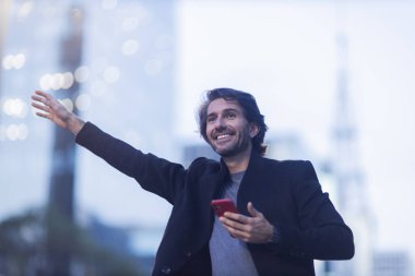 Alacakaranlıkta akıllı telefonlu genç bir adamın görüntüsü. Arka planda şehir manzarası olan bir taksi çağırıyor. Yüksek kalite fotoğraf. 