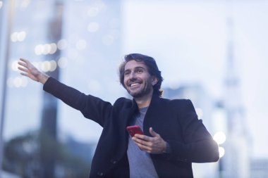 Alacakaranlıkta akıllı telefonlu genç bir adamın görüntüsü. Arka planda şehir manzarası olan bir taksi çağırıyor. Yüksek kalite fotoğraf. 