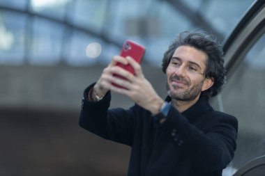 Alacakaranlıkta arka planda bulanık bir manzara olan kapalı bir alanda akıllı telefon kullanan genç bir adamın görüntüsü. Yüksek kalite fotoğraf. Telefondan selfie çekiyorum. Video görüşmesi yapıyorum. Alacakaranlıkta akıllı telefon kullanan genç bir adamın görüntüsü. 