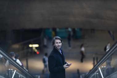 Alacakaranlıkta arka planda bulanık bir manzara olan kapalı bir alanda akıllı telefon kullanan genç bir adamın görüntüsü. Yüksek kalite fotoğraf. Telefonda mesajlaşıyoruz. Gece vakti şehir manzaralı akıllı telefon kullanan genç bir adamın görüntüsü.