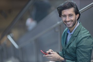 Arka planda bulanık bir manzara olan kapalı alanda akıllı telefon kullanan genç bir adamın görüntüsü. Yüksek kalite fotoğraf. Telefondaki mesajlaşma.. 