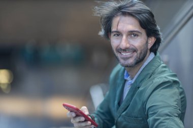 Arka planda bulanık bir manzara olan kapalı alanda akıllı telefon kullanan genç bir adamın görüntüsü. Yüksek kalite fotoğraf. Telefondaki mesajlaşma.. 
