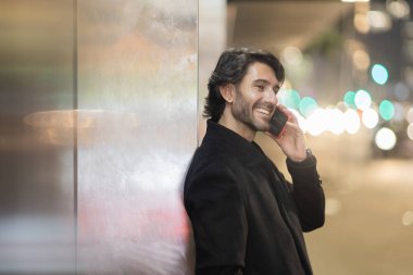 Gece vakti arka planda şehir manzarası olan akıllı telefon kullanan genç bir adamın görüntüsü. Yüksek kalite fotoğraf. Gece vakti arka planda şehir manzarası olan akıllı telefon kullanan genç bir adamın görüntüsü. Yüksek kalite fotoğraf.