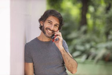 Şehirdeki yeşil bir parkta gündüz vakti akıllı telefonlu genç bir adam görülüyor..