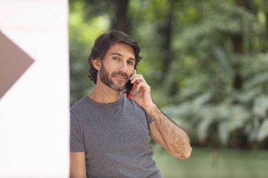 Şehirdeki yeşil bir parkta gündüz vakti akıllı telefonlu genç bir adam görülüyor..
