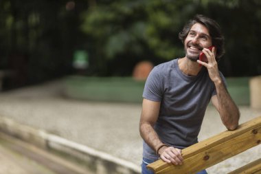 Şehirdeki yeşil bir parkta gündüz vakti akıllı telefonlu genç bir adam görülüyor..