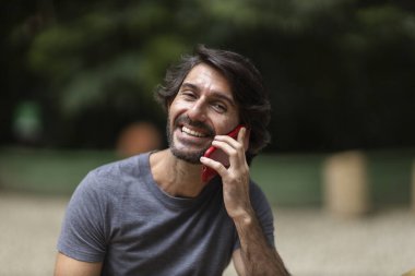 Şehirdeki yeşil bir parkta gündüz vakti akıllı telefonlu genç bir adam görülüyor..