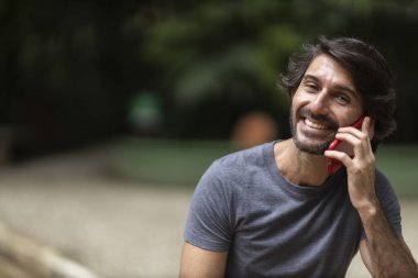 Şehirdeki yeşil bir parkta gündüz vakti akıllı telefonlu genç bir adam görülüyor..