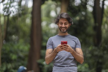 Şehirdeki yeşil bir parkta gündüz vakti akıllı telefonlu genç bir adam görülüyor..