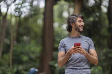 Şehirdeki yeşil bir parkta gündüz vakti akıllı telefonlu genç bir adam görülüyor..