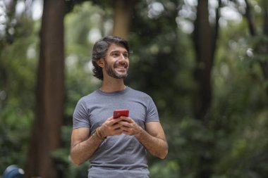 Şehirdeki yeşil bir parkta gündüz vakti akıllı telefonlu genç bir adam görülüyor..