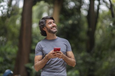 Şehirdeki yeşil bir parkta gündüz vakti akıllı telefonlu genç bir adam görülüyor..