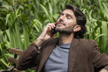 Gündüz vakti akıllı telefonlu genç bir adamın şehirdeki yeşil bir parktaki bankta oturduğunu görmek. Yüksek kalite fotoğraf.