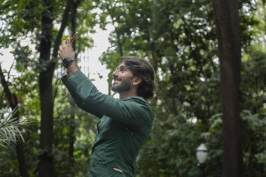 Şehirdeki yeşil bir parkta gündüz vakti akıllı telefonlu genç bir adam görülüyor..