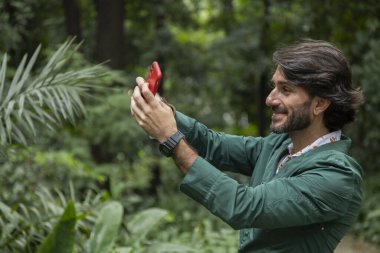 Şehirdeki yeşil bir parkta gündüz vakti akıllı telefonlu genç bir adam görülüyor..