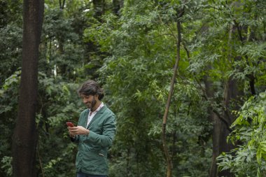 Şehirdeki yeşil bir parkta gündüz vakti akıllı telefonlu genç bir adam görülüyor..