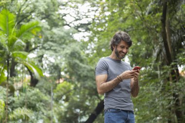 Şehirdeki yeşil bir parkta gündüz vakti akıllı telefonlu genç bir adam görülüyor..