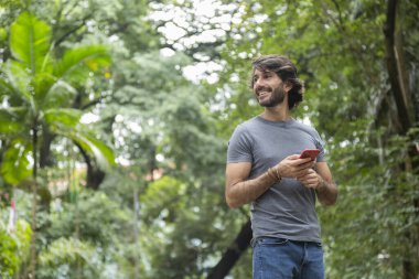 Şehirdeki yeşil bir parkta gündüz vakti akıllı telefonlu genç bir adam görülüyor..