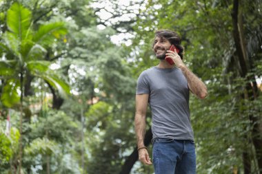 Şehirdeki yeşil bir parkta gündüz vakti akıllı telefonlu genç bir adam görülüyor..