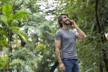 Şehirdeki yeşil bir parkta gündüz vakti akıllı telefonlu genç bir adam görülüyor..