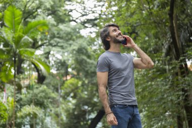 Şehirdeki yeşil bir parkta gündüz vakti akıllı telefonlu genç bir adam görülüyor..