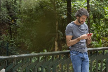 Şehirdeki yeşil bir parkta gündüz vakti akıllı telefonlu genç bir adam görülüyor..