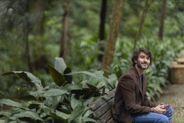 Gündüz vakti akıllı telefonlu genç bir adamın şehirdeki yeşil bir parktaki bankta oturduğunu görmek. Yüksek kalite fotoğraf.