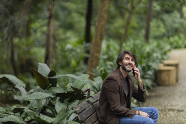 Gündüz vakti akıllı telefonlu genç bir adamın şehirdeki yeşil bir parktaki bankta oturduğunu görmek. Yüksek kalite fotoğraf.