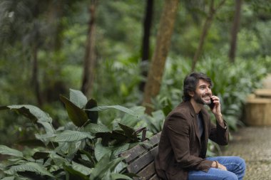 Gündüz vakti akıllı telefonlu genç bir adamın şehirdeki yeşil bir parktaki bankta oturduğunu görmek. Yüksek kalite fotoğraf.