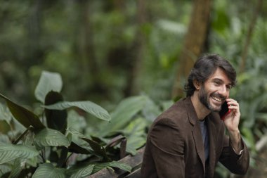 Gündüz vakti akıllı telefonlu genç bir adamın şehirdeki yeşil bir parktaki bankta oturduğunu görmek. Yüksek kalite fotoğraf.