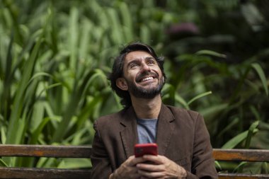 Gündüz vakti akıllı telefonlu genç bir adamın şehirdeki yeşil bir parktaki bankta oturduğunu görmek. Yüksek kalite fotoğraf.