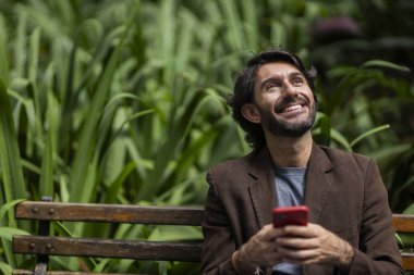 Gündüz vakti akıllı telefonlu genç bir adamın şehirdeki yeşil bir parktaki bankta oturduğunu görmek. Yüksek kalite fotoğraf.