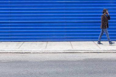 Akıllı telefonlu genç bir adamın gündüz vakti mavi bir geçmişle sokakta yürüdüğünü görmek. Yüksek kalite fotoğraf.