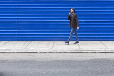 Akıllı telefonlu genç bir adamın gündüz vakti mavi bir geçmişle sokakta yürüdüğünü görmek. Yüksek kalite fotoğraf.