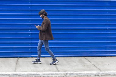 Akıllı telefonlu genç bir adamın gündüz vakti mavi bir geçmişle sokakta yürüdüğünü görmek. Yüksek kalite fotoğraf.