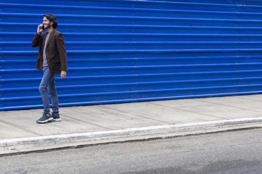 Akıllı telefonlu genç bir adamın gündüz vakti mavi bir geçmişle sokakta yürüdüğünü görmek. Yüksek kalite fotoğraf.