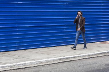 Akıllı telefonlu genç bir adamın gündüz vakti mavi bir geçmişle sokakta yürüdüğünü görmek. Yüksek kalite fotoğraf.