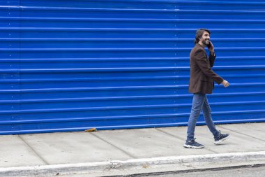 Akıllı telefonlu genç bir adamın gündüz vakti mavi bir geçmişle sokakta yürüdüğünü görmek. Yüksek kalite fotoğraf.
