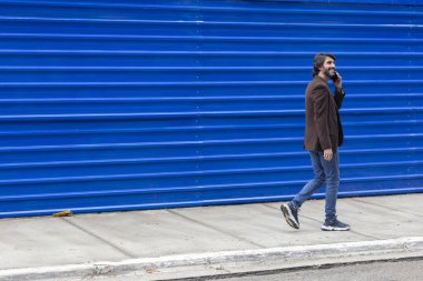 Akıllı telefonlu genç bir adamın gündüz vakti mavi bir geçmişle sokakta yürüdüğünü görmek. Yüksek kalite fotoğraf.