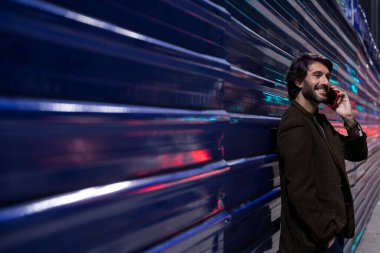 Gece vakti akıllı telefon kullanan genç bir adam şehir ışıklarının olduğu mavi bir duvara yaslanıyor. Gece vakti akıllı telefon kullanan genç adam, arka planda şehir manzarası olan mavi bir duvara yaslanıyor.. 