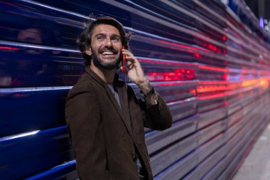 Gece vakti akıllı telefon kullanan genç bir adam şehir ışıklarının olduğu mavi bir duvara yaslanıyor. Gece vakti akıllı telefon kullanan genç adam, arka planda şehir manzarası olan mavi bir duvara yaslanıyor.. 